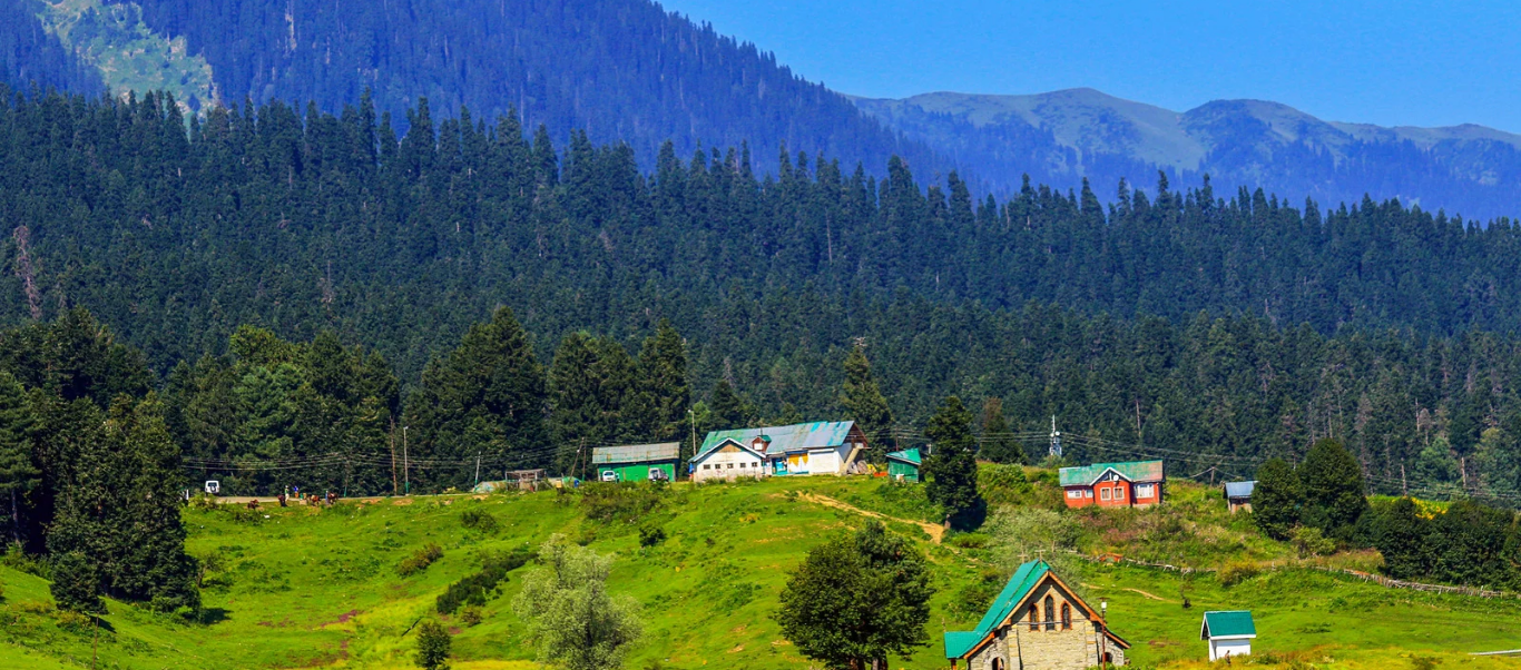 Wide-angle view of the scenic hill station Katra in Jammu, featuring a dense cluster of white buildings nestled at the base of lush, rolling green mountains under a misty sky.