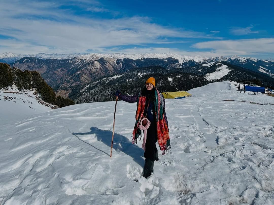 A traveler standing in deep snow during the Raghupur Fort trek near Jibhi, with a panoramic view of the snow-capped Himalayan mountain ranges in the background.
