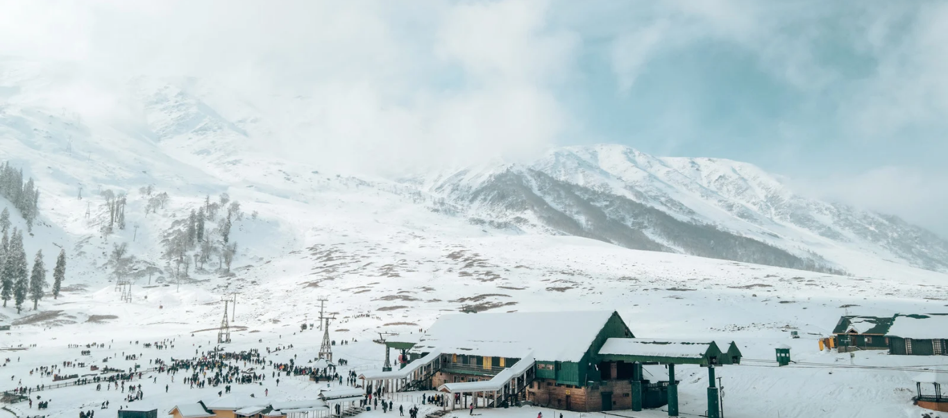 A snow-covered mountain resort in Srinagar featuring green-roofed buildings and ski lift infrastructure against a backdrop of towering, mist-shrouded Himalayan peaks.